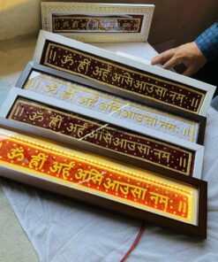 Framed decorative panels with gold-stitched Om symbols on red and white backgrounds, arranged in a diagonal display, a hand nearby wearing a ring.