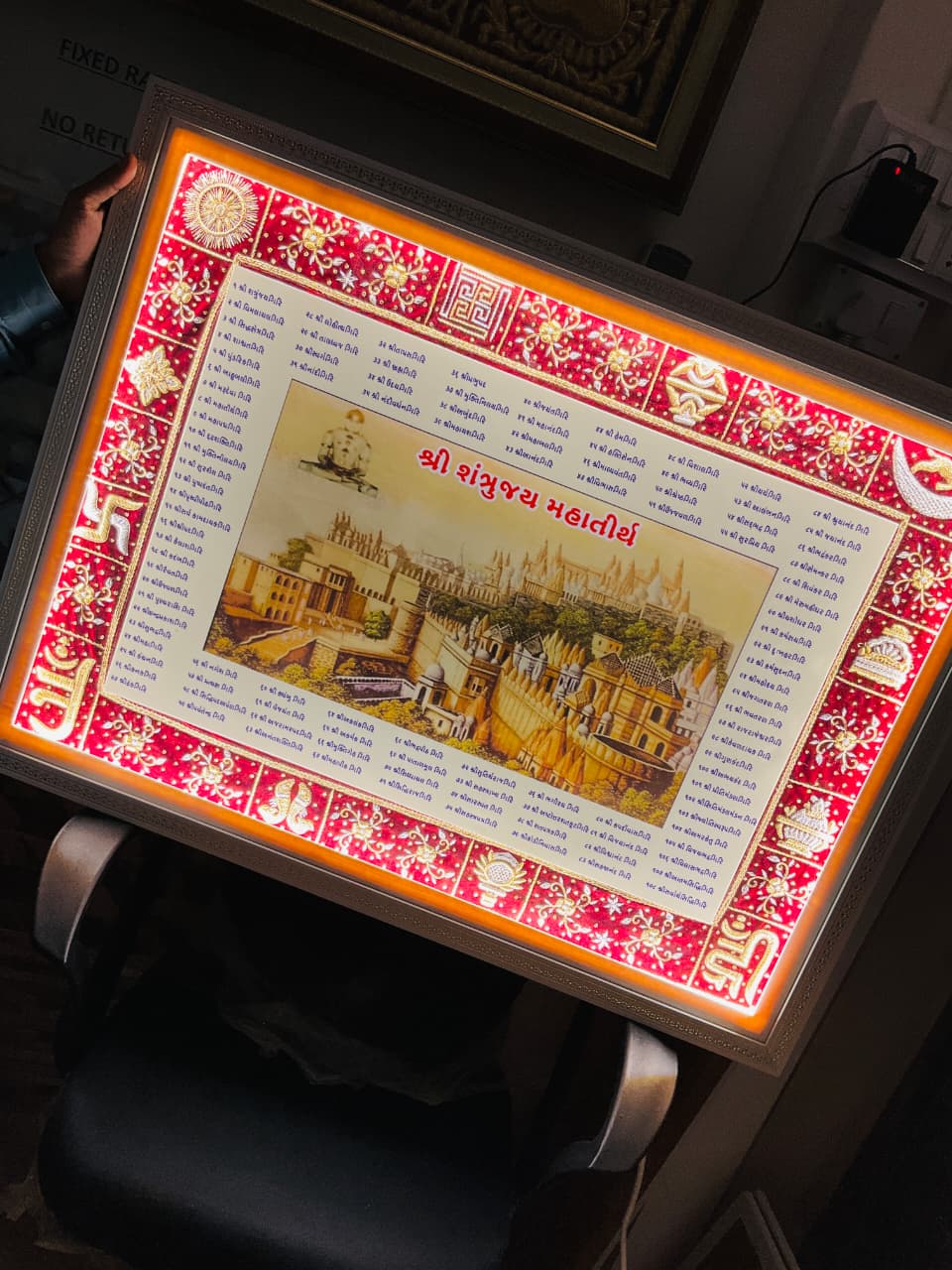 Backlit ornate framed poster with a red and gold border, featuring Gujarati text and a cityscape illustration.