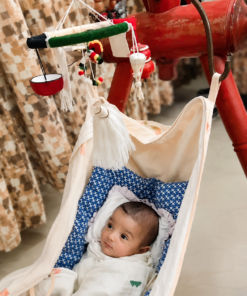 Baby sleeping in a fabric baby hammock suspended from a red frame, with colorful toys and tassels above.