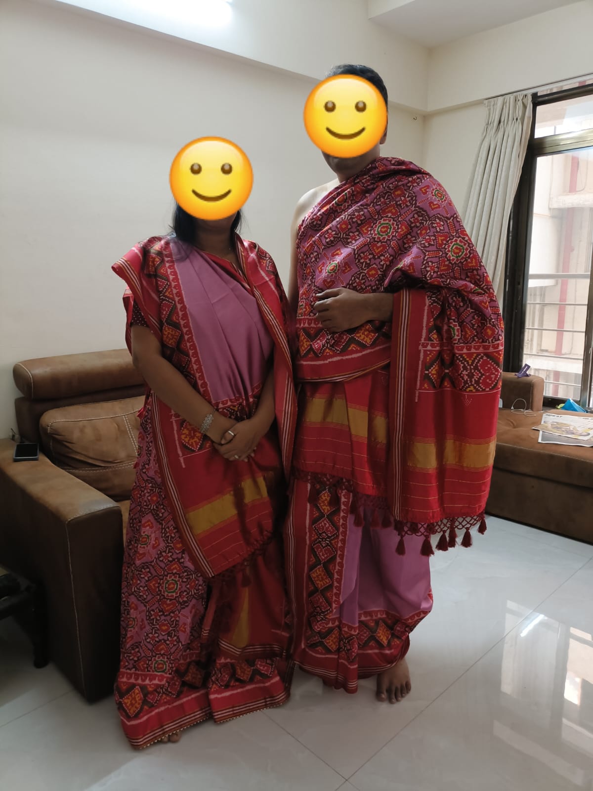 Two people stand side by side in a living room, wearing matching pink and red traditional sarees with patterned borders.