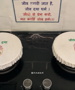 Two white round cake covers on a black stovetop, each showing green Hindi text 'जीव द्या' above a colored hand symbol with a small emblem in the palm.