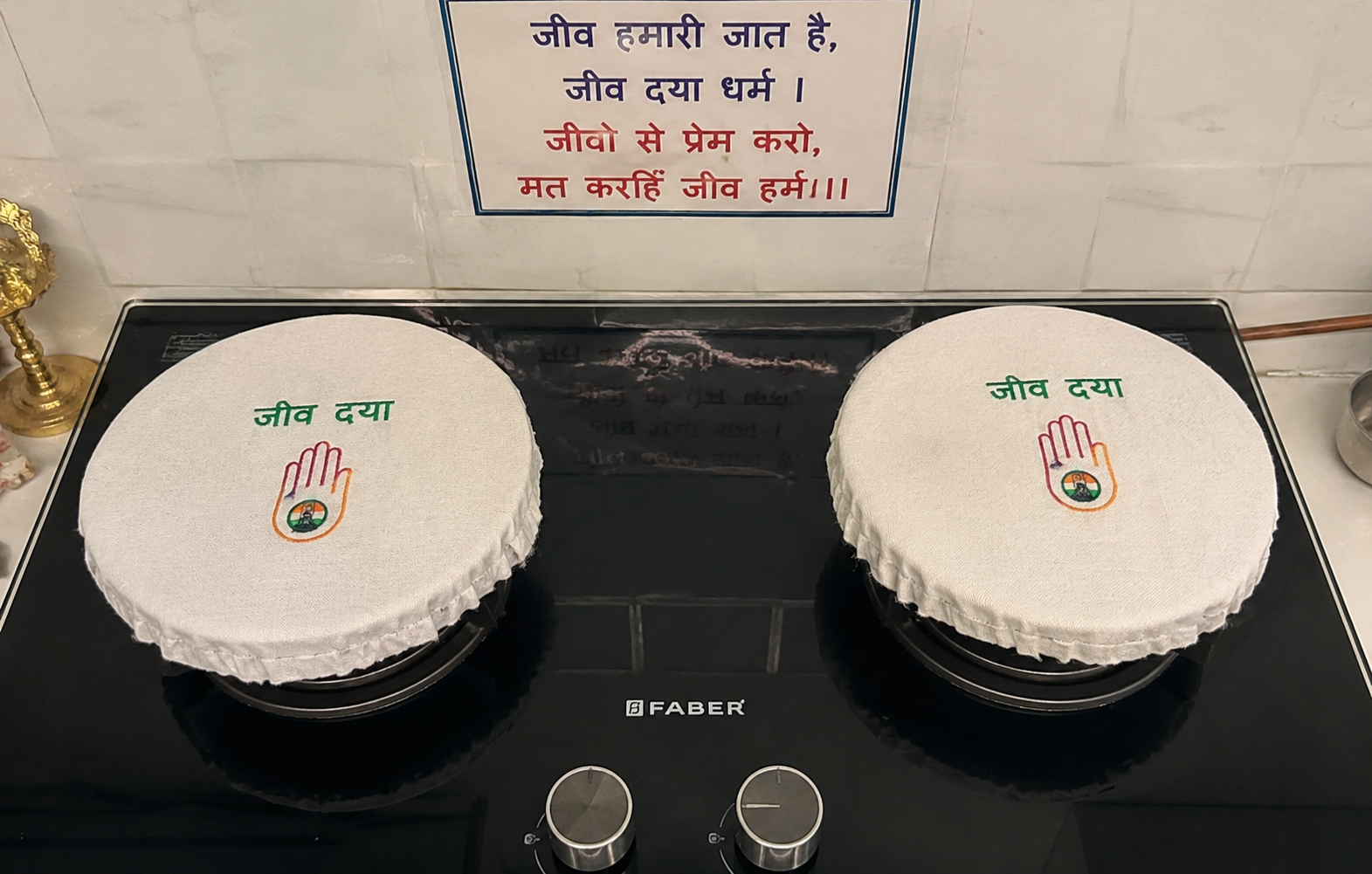 Two white round cake covers on a black stovetop, each showing green Hindi text 'जीव द्या' above a colored hand symbol with a small emblem in the palm.
