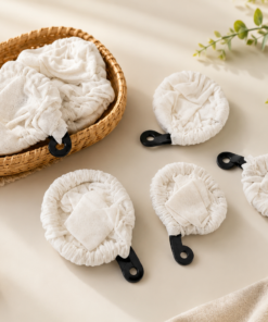 Wicker basket holding a soft white cloth, with five round fabric facial rounds with black looping handles laid out on a beige surface nearby.