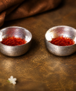 Two silver bowls filled with red saffron threads on a brown textured surface, with small white flowers nearby.