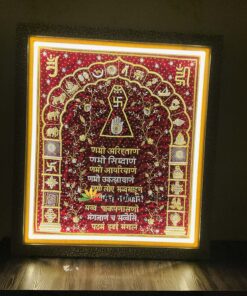 Framed red embroidered Hindu ceremonial textile with gold motifs and Sanskrit text, lit from above, featuring sacred symbols like a central swastika and geometric designs.
