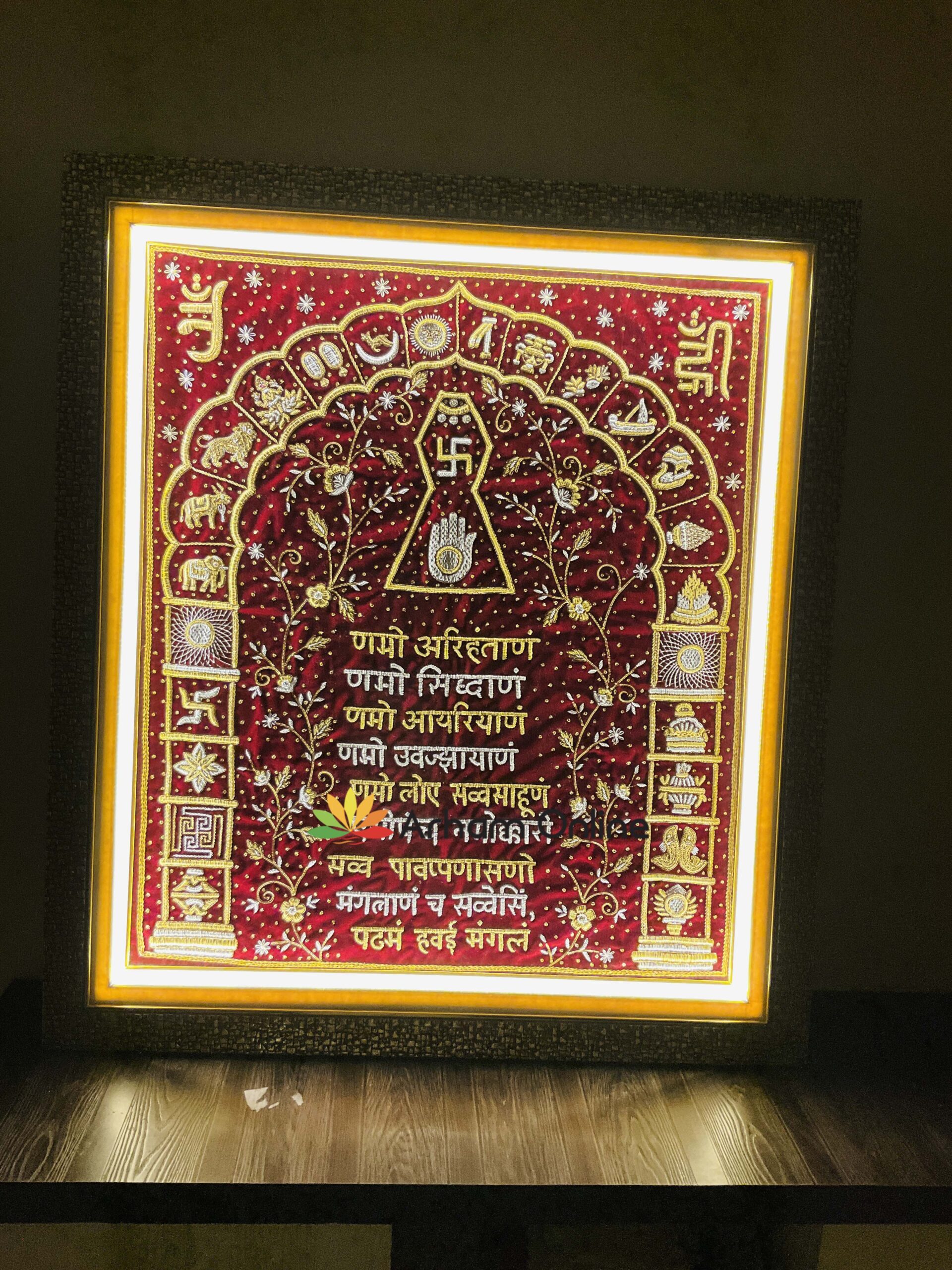Framed red embroidered Hindu ceremonial textile with gold motifs and Sanskrit text, lit from above, featuring sacred symbols like a central swastika and geometric designs.