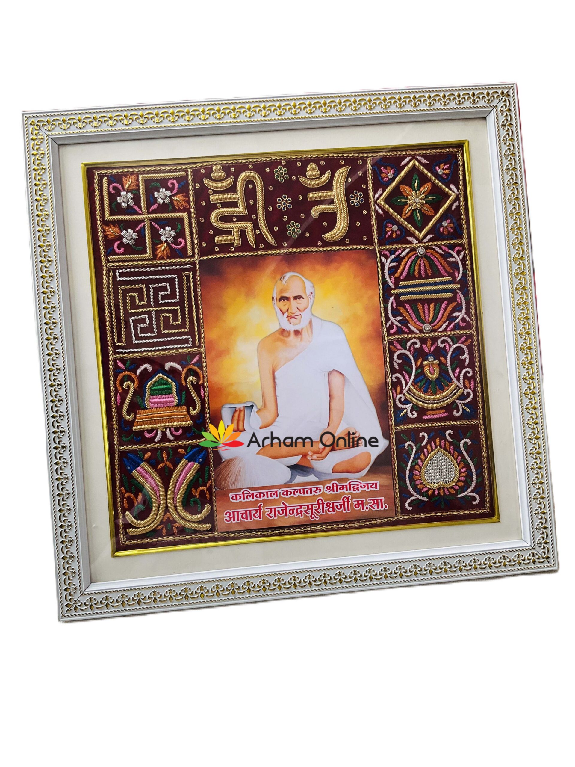 Decorative framed devotional artwork with a central portrait of a sage in white robes, surrounded by embroidered panels and a gold-trimmed frame.