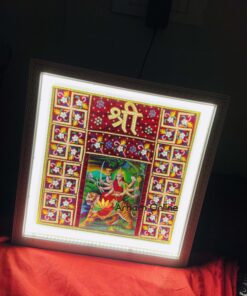 Illuminated framed Hindu artwork depicting Durga in the center, surrounded by colorful floral tile borders, resting on red fabric.