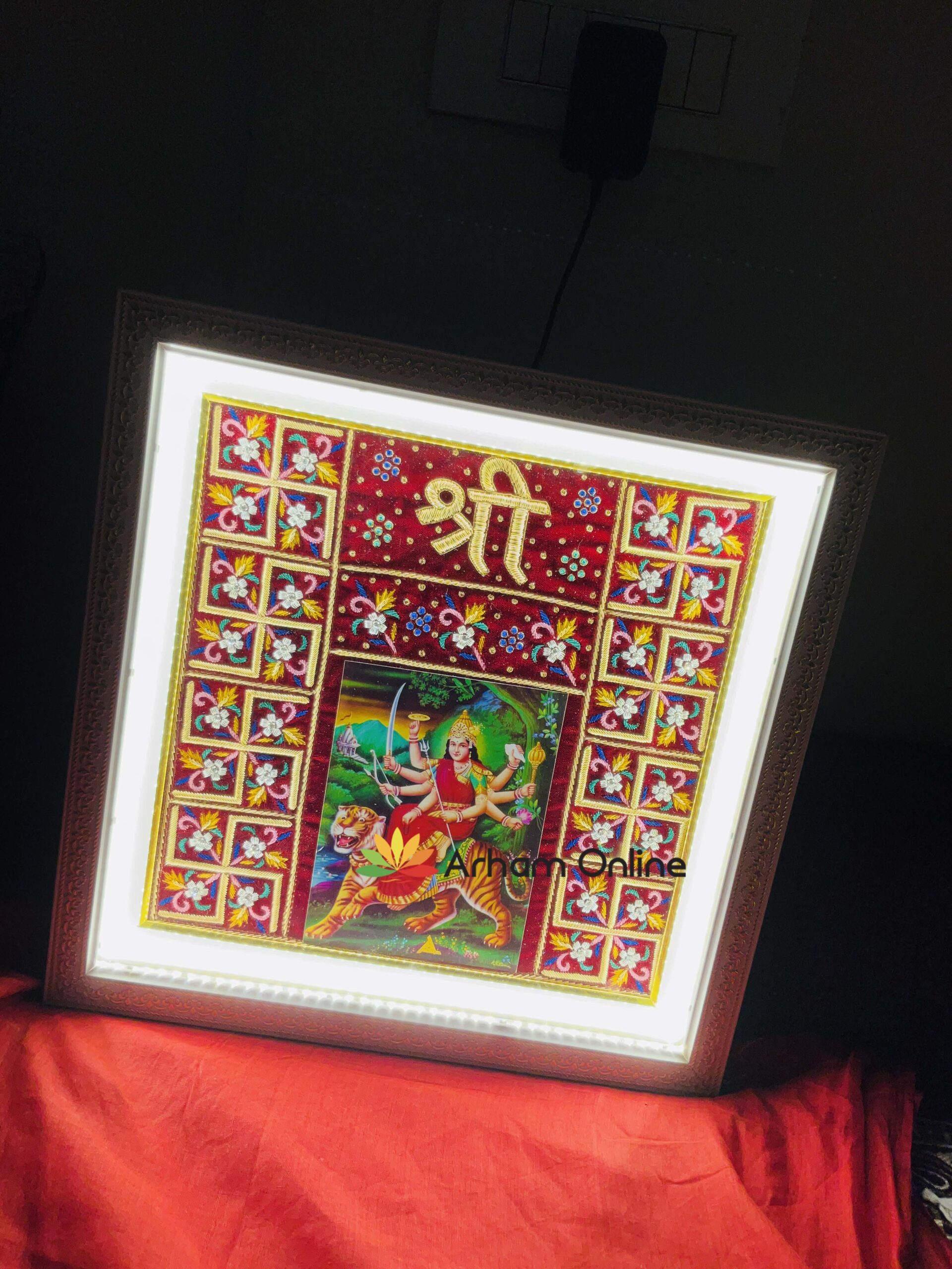Illuminated framed Hindu artwork depicting Durga in the center, surrounded by colorful floral tile borders, resting on red fabric.
