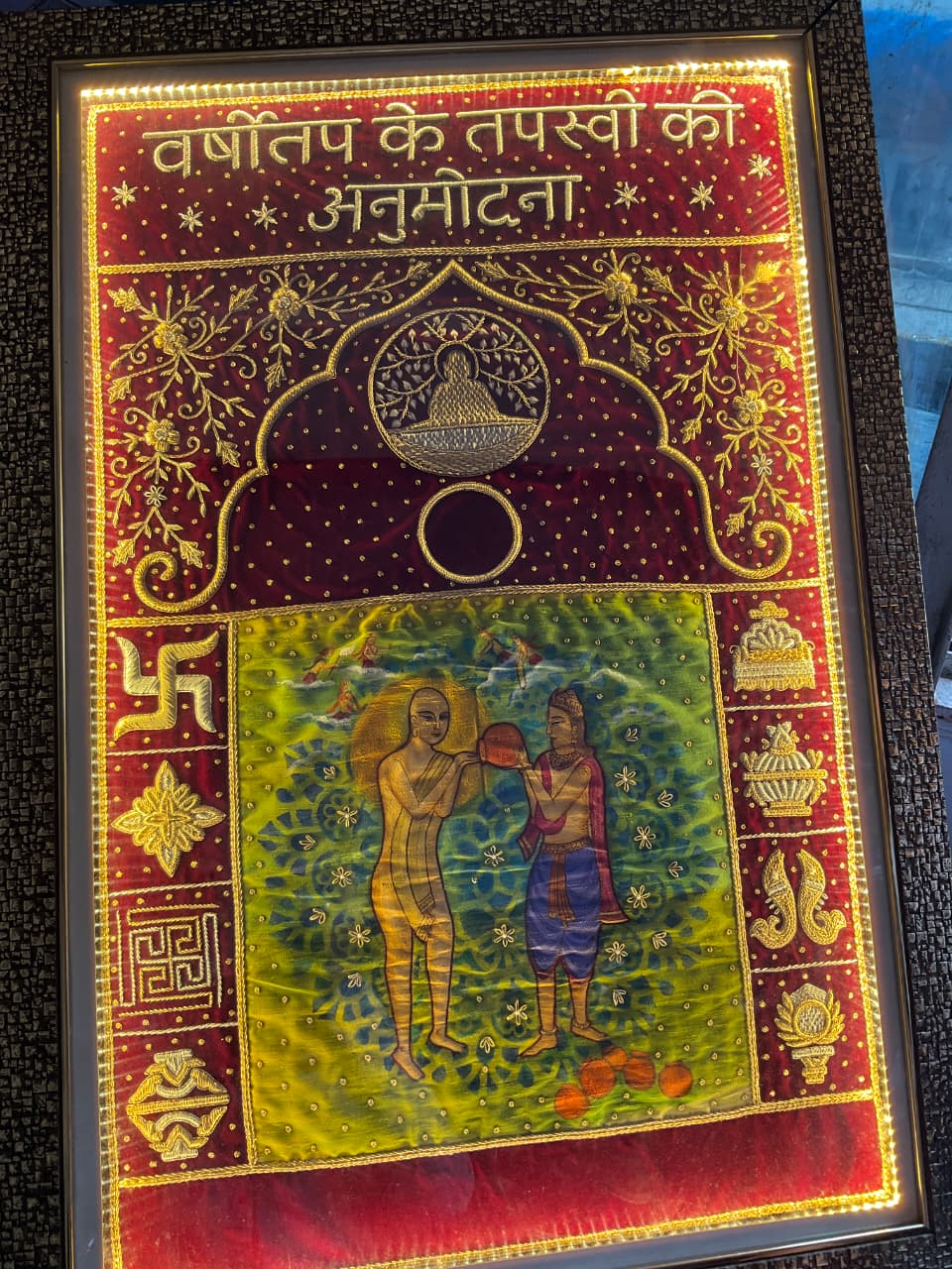 Framed embroidered textile with gold floral border and Hindi text at the top; central arch motif and a colorful scene of two figures exchanging an object in a green field, surrounded by ornate decorations.