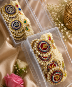 Two ornate pearl- and gold-embellished jewelry pieces in clear plastic boxes on satin fabric, with a pink rose nearby.