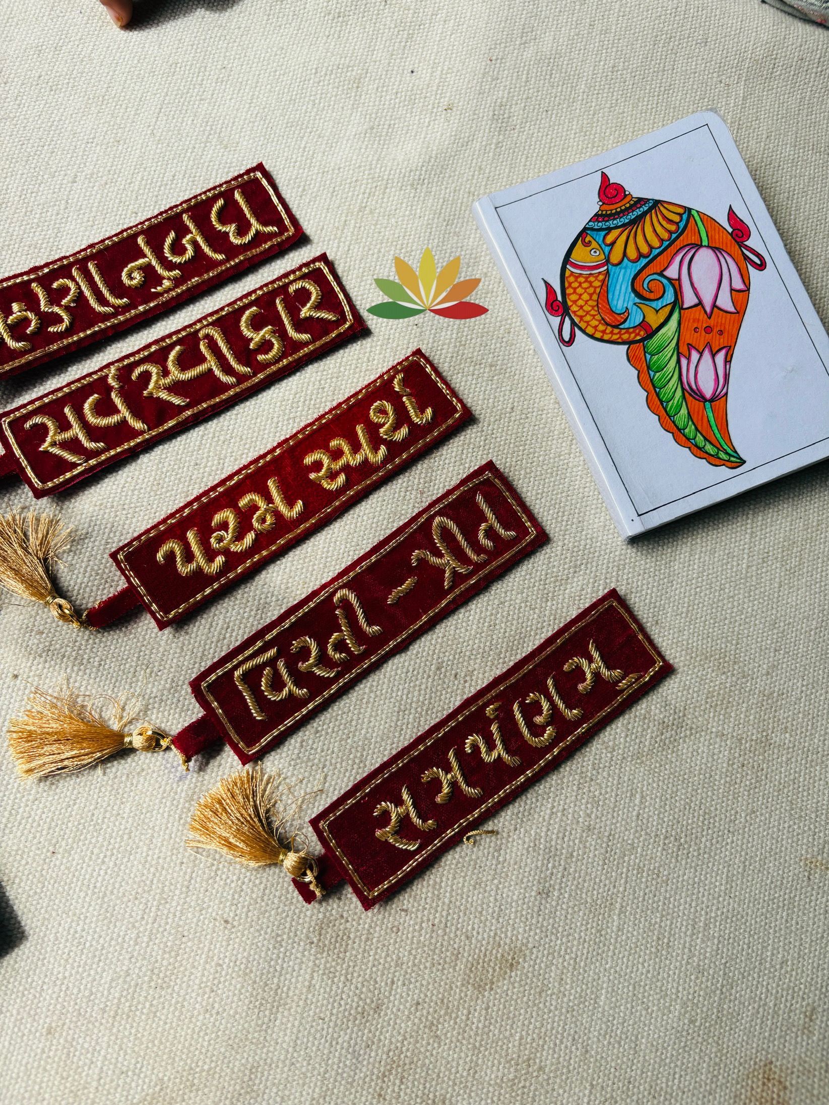 handwork book mark Red velvet decorative banners with gold embroidery and tassels on a beige fabric, next to a colorful elephant illustration on a card.
