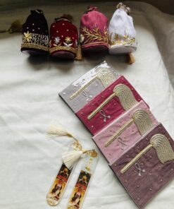 Four ornate velvet pouches (black, red, pink, white) with gold embroidery and tassels laid on a light fabric, with decorative booklets nearby.