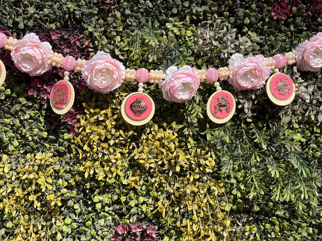 Decorative garland with large pink paper roses and pink beaded ornaments against a lush green leafy backdrop.
