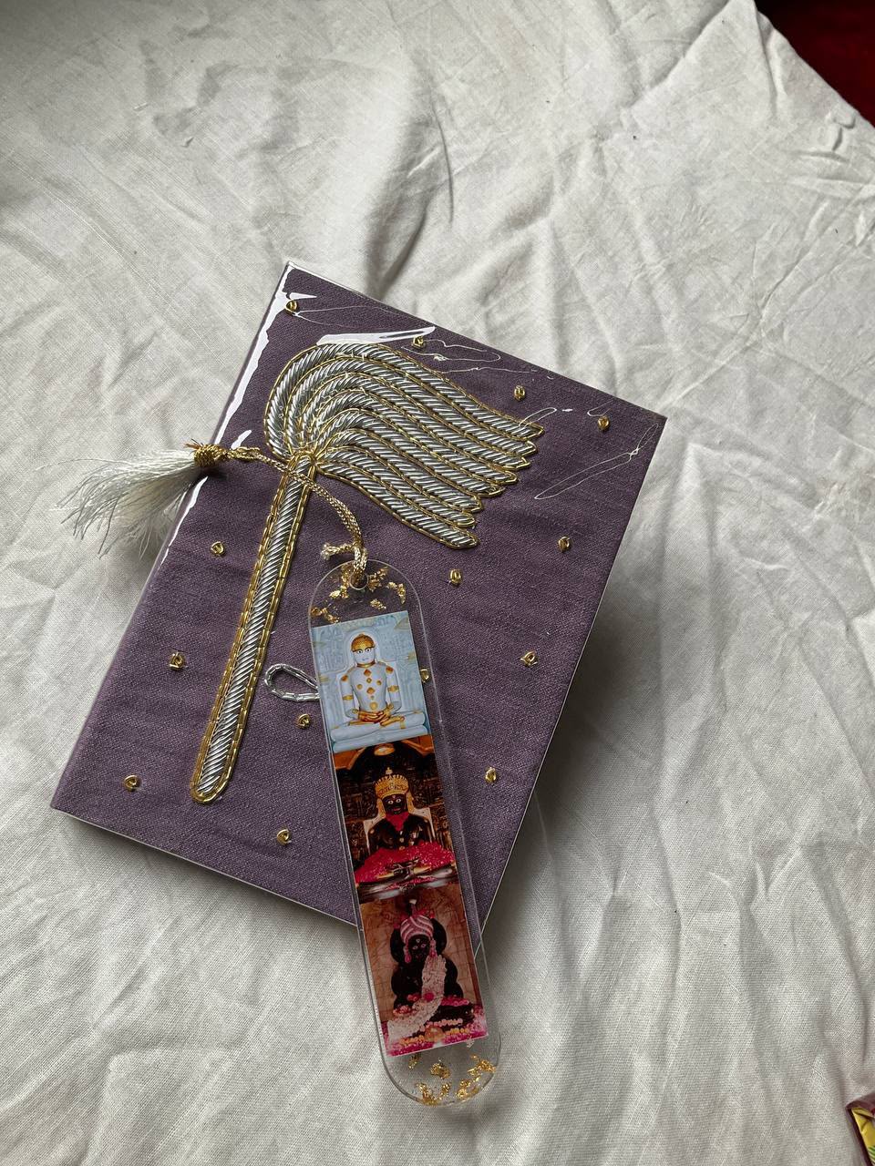 WhatsApp Image 2026-07-03 at 5.00.08 PM (1) Purple gift box adorned with a gold braided palm-leaf ornament and white tassel, with a vertical, multi-image bookmark resting on it.