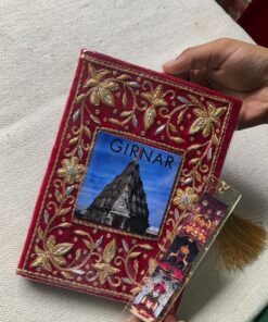 Ornately embroidered red cover with gold floral patterns, featuring a temple photo labeled GIRNAR and a decorative bookmark hanging on the side, held by a hand.
