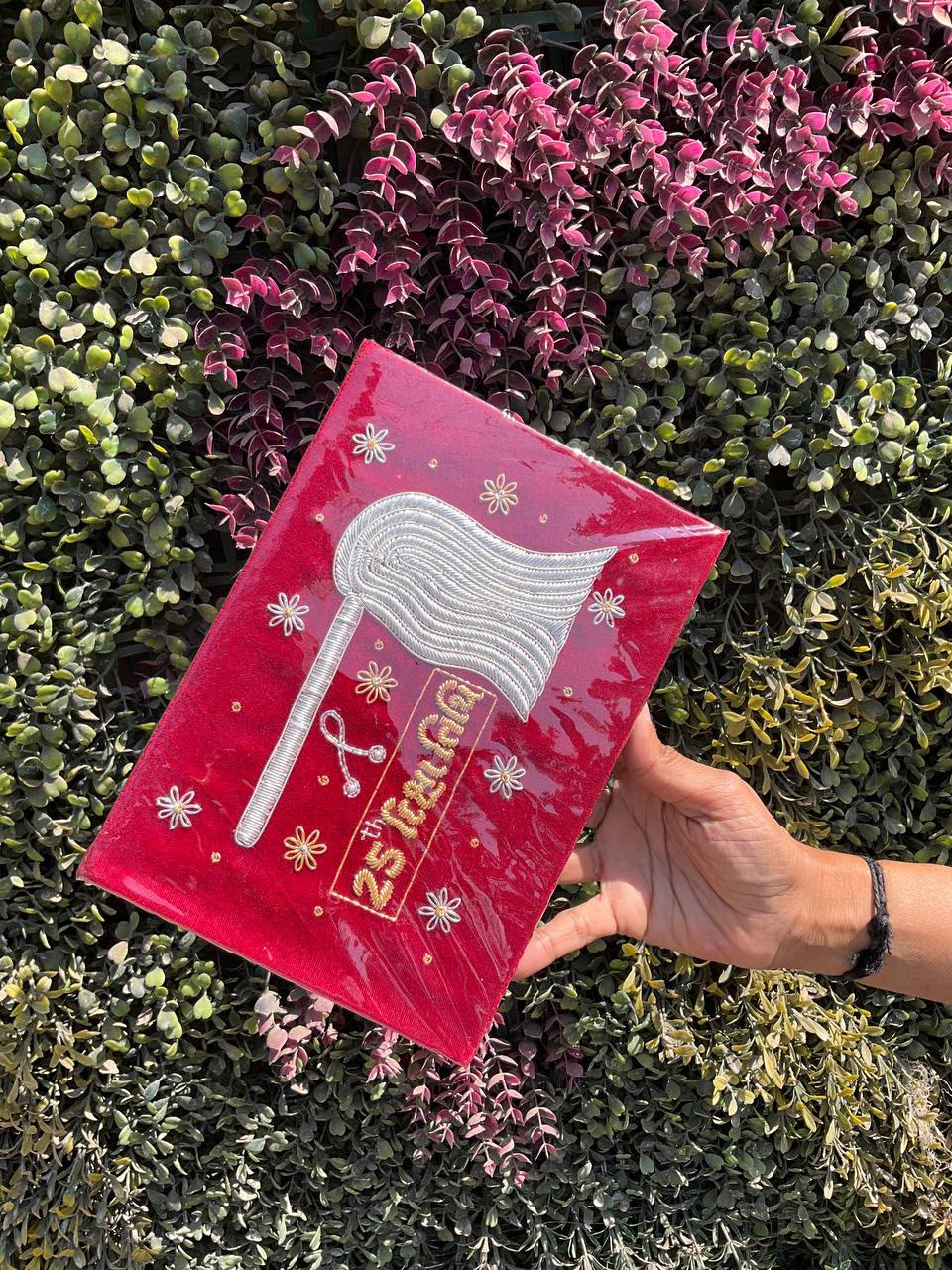 A red graduation cap held in a hand, featuring a silver axe emblem and small white flower decorations against a leafy green background.