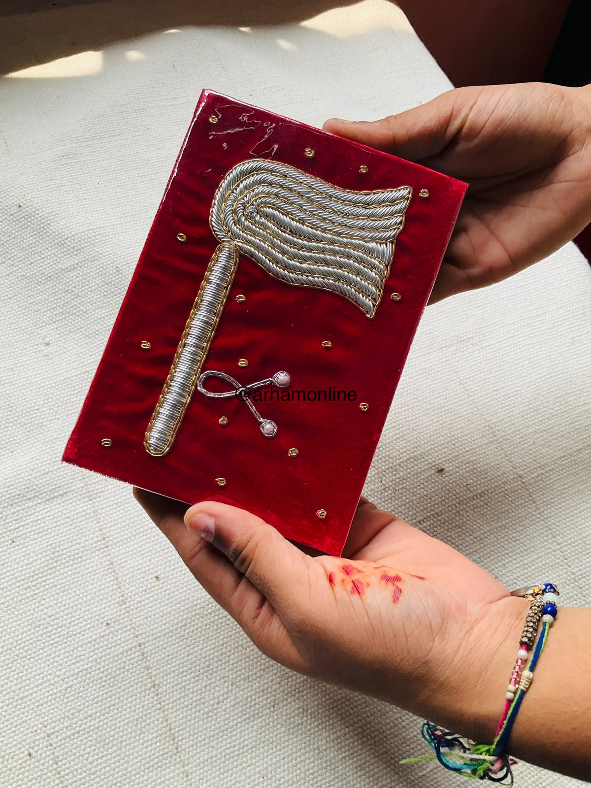 WhatsApp Image 2026-07-03 at 5.00.09 PM (5) Hand holding a red plaque with a raised silver rope-wrapped axe design and small gold studs dotted around the background