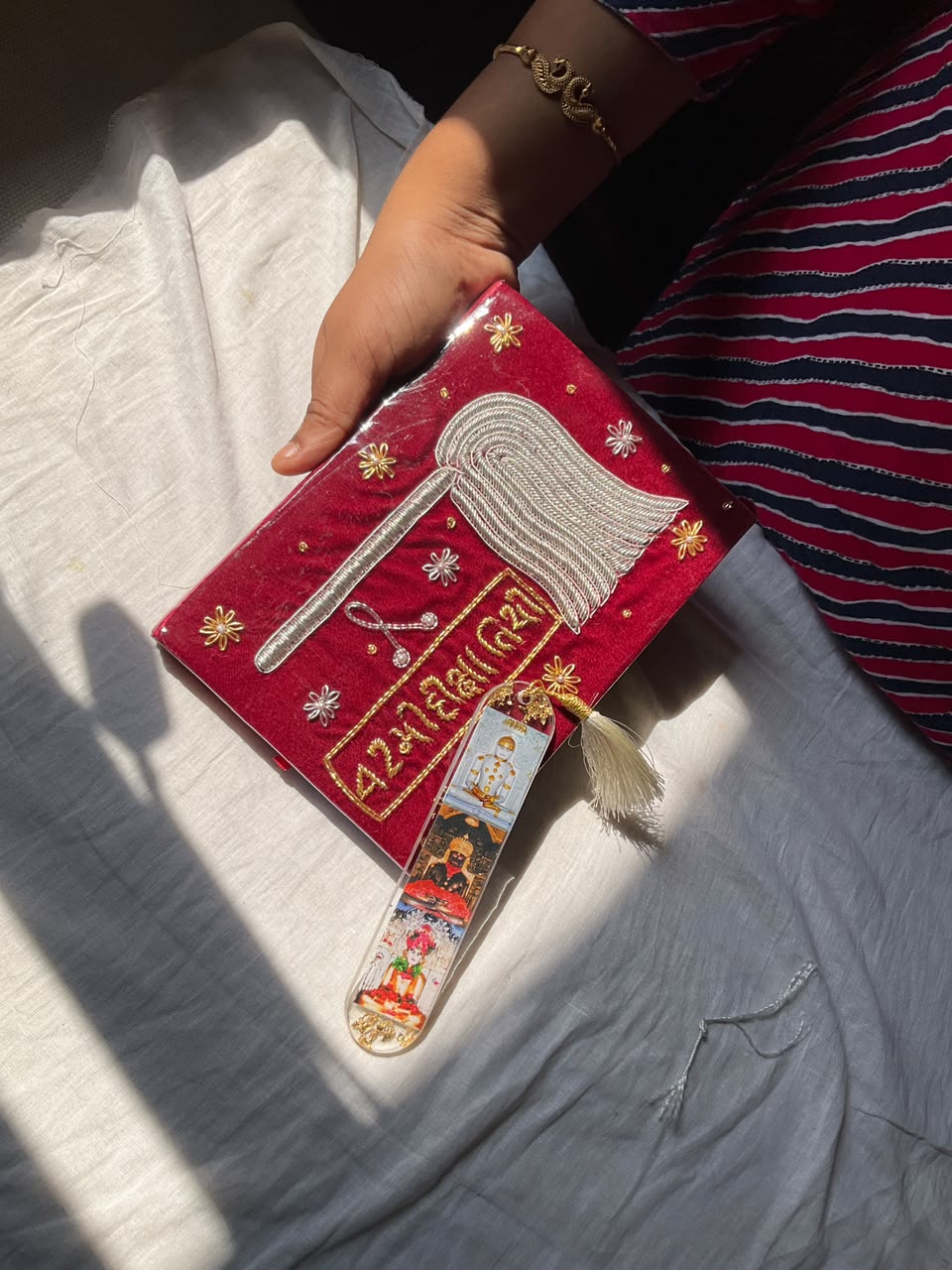 WhatsApp Image 2026-07-03 at 5.00.09 PM (7) Red velvet devotional pouch with silver embroidery and a tassel, held by a person; a laminated strip of religious images attached.