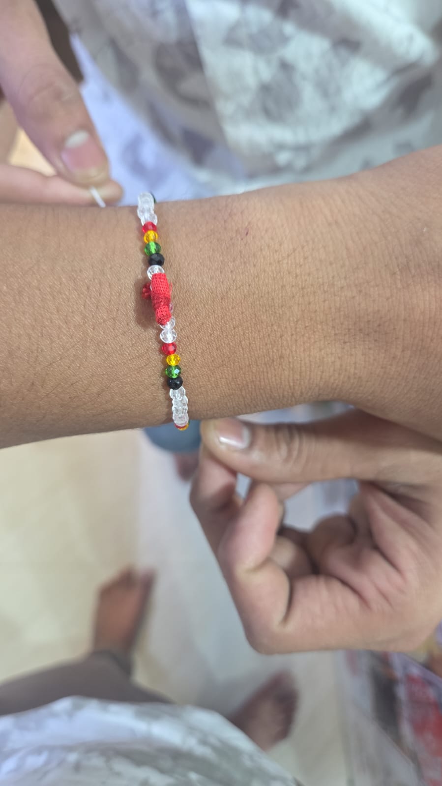 WhatsApp Image 2026-07-17 at 11.16.25 AM Beaded bracelet on wrist with red fabric accent and bright colored beads (red, yellow, green, black) being adjusted by hand
