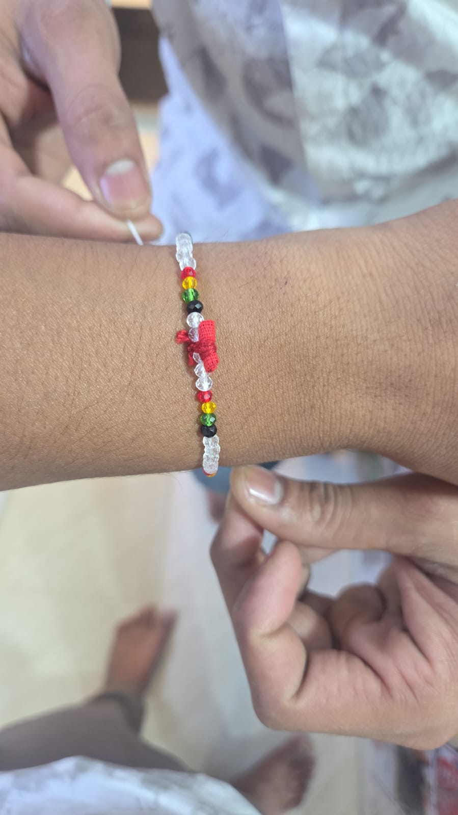 WhatsApp Image 2026-07-17 at 11.19.22 AM Beaded bracelet on wrist with white translucent beads and a row of multicolored beads (red, yellow, green, black) and red knotting thread in the center.