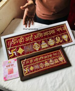 Framed red and gold embroidered strips with Sanskrit mantras and symbols, displayed on a white table with a perched hand nearby.