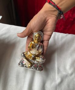 Hand holding a small gold-and-silver Buddha statue in a meditative pose, worn with a red-and-black thread bracelet and white fabric background.